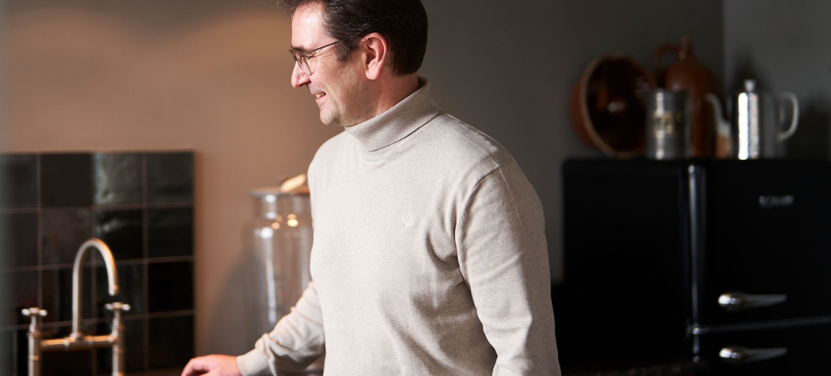 Ein Mann in einem hellen Pullover vor einem hochwertigem alten Waschebecken, Das Waschbecken ist Kupferfarben und die Küche ist schwarz gehalten.