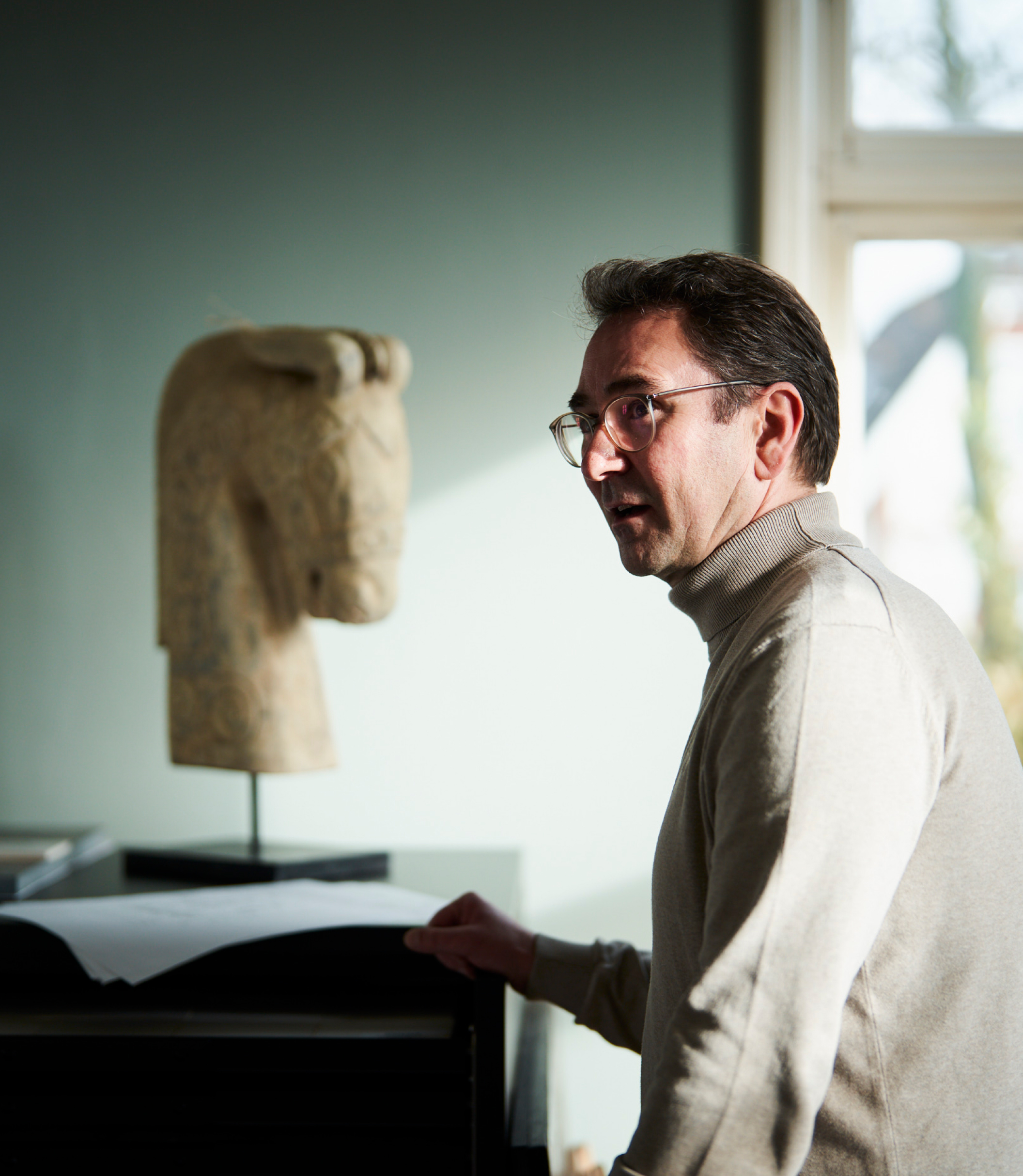 Ein Mann mit einem hellen Pullover vor einem schwarz lackiertem Sekretär. Auf dem Sekretär steht eine Skulptur von einem Pferdekop. Im Hintergrund erkennt man Baumaterialien und eine Terrassentür.