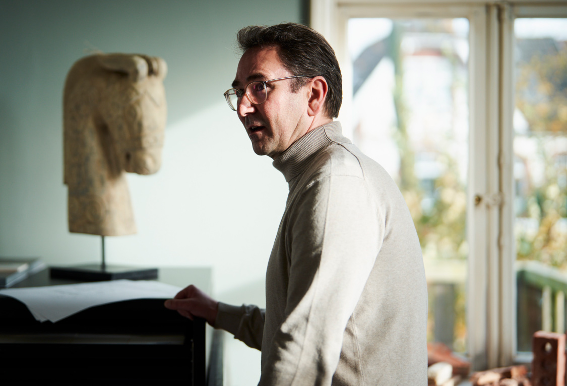 Ein Mann mit einem hellen Pullover vor einem schwarz lackiertem Sekretär. Auf dem Sekretär steht eine Skulptur von einem Pferdekop. Im Hintergrund erkennt man Baumaterialien und eine Terrassentür.