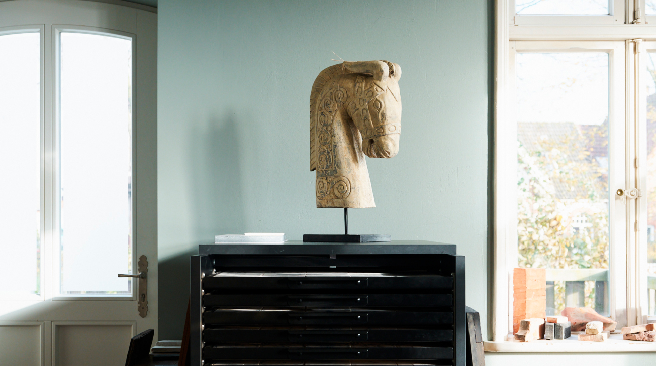 Zwischen einem Fenster und einer Tür mit Sichtfenstern steht eine schwarze Anrichte. Auf dieser Anrichte steht ein sandfarbener Pferdekopf.