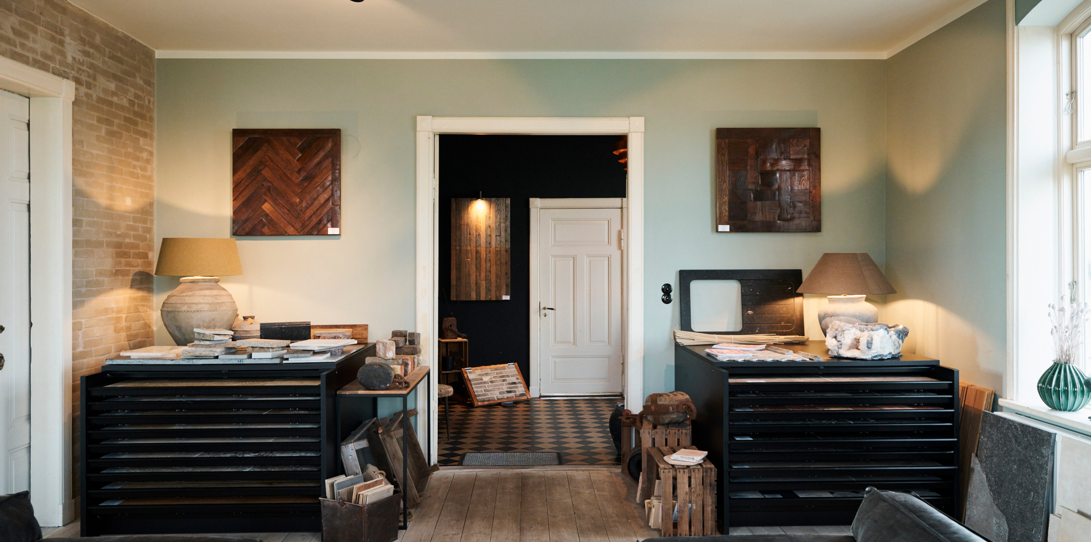 Ansicht von einem großen quadratischen Raum auf die Zimmertür. Rechts und Links von der Tür stehen schwarze Anrichten auf denen jeweils eine beige Lampe und Materialien liegen. Über den Anrichten hängen dunkle Bilder. Durch die Zimmertür kann man in den Flur gucken und sieht eine weitere Tür, diese ist geschlossen.